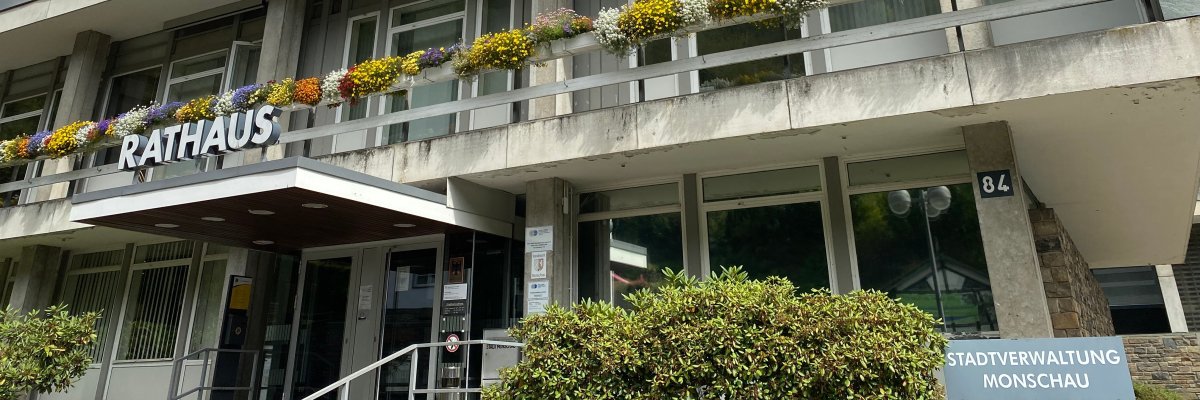 Das Bild zeigt das Monschauer Rathaus. Ein großes Gebäude mit heller Fassade und Blumen in den Kästen auf dem Balkon.