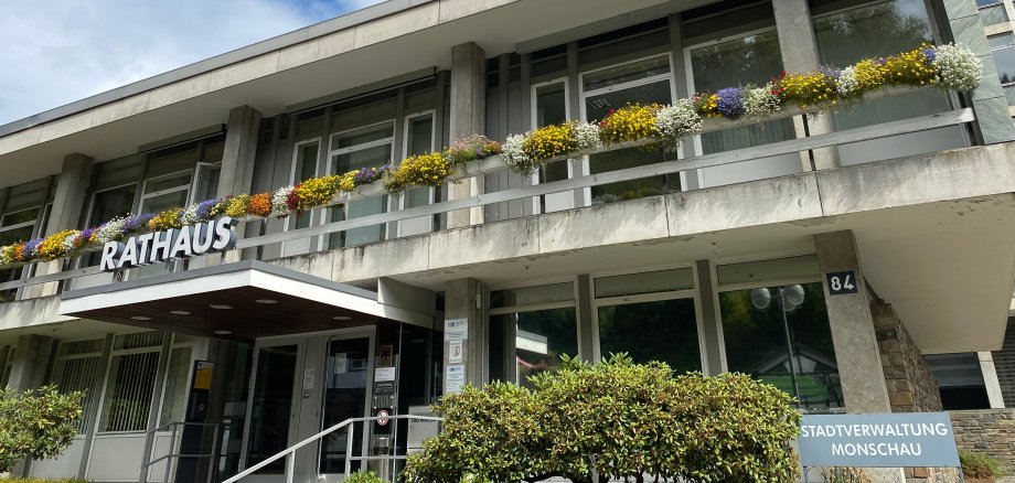 Das Bild zeigt das Monschauer Rathaus. Ein großes Gebäude mit heller Fassade und Blumen in den Kästen auf dem Balkon.